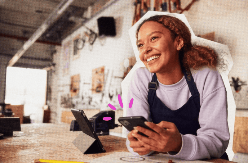 female presenting solopreneur with apronat work counter with card reader holding phone smiling